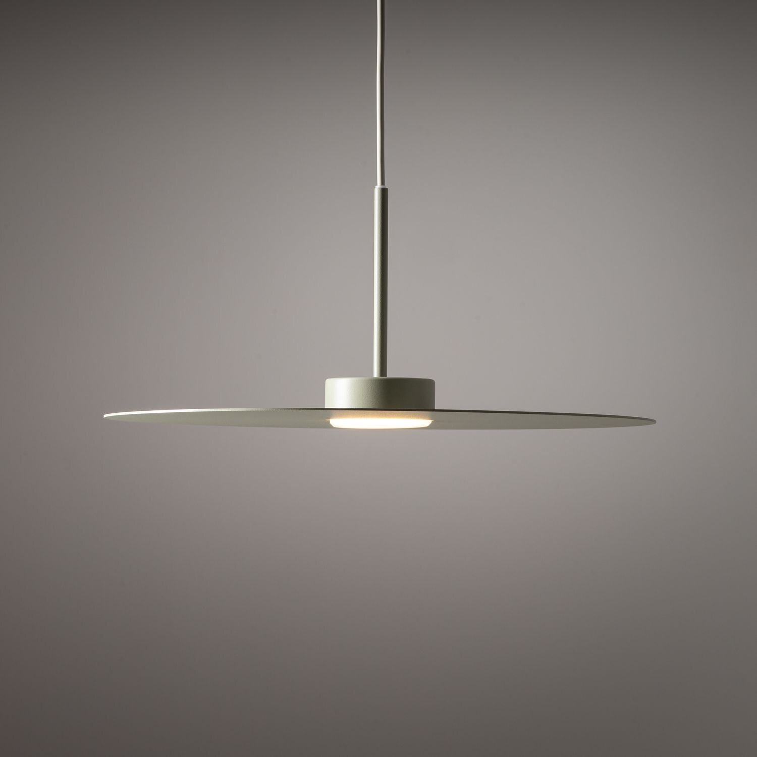 Lampa wisząca o minimalistycznym designie z płaskim, okrągłym kloszem w kolorze szarym. Lampa jest zawieszona na cienkim przewodzie, a światło emituje z centralnej części klosza.