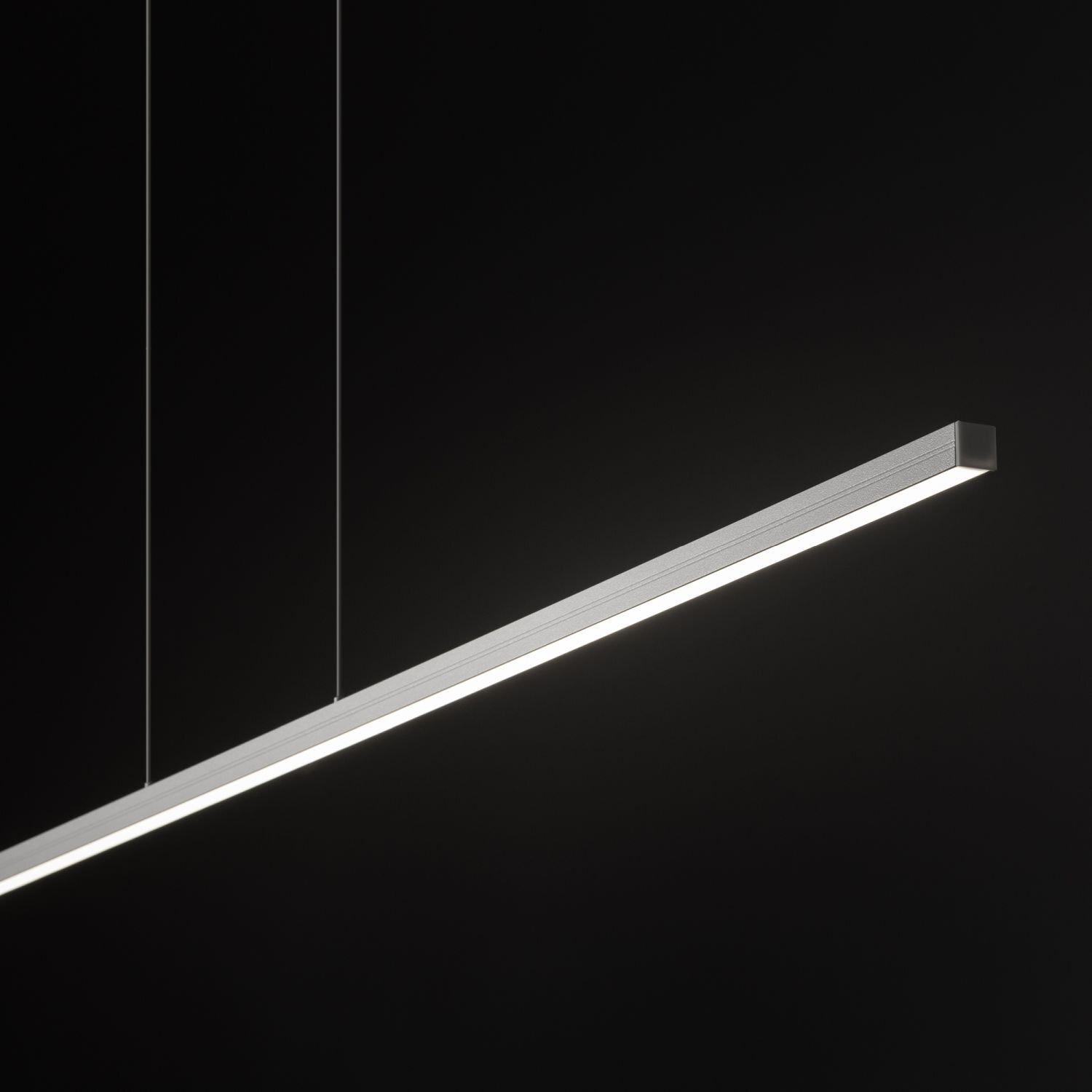Na obrazie widoczna jest nowoczesna lampa wisząca LED o prostokątnym kształcie, zawieszona na cienkich linkach. Tło jest ciemne, co podkreśla jasność emitowanego światła.