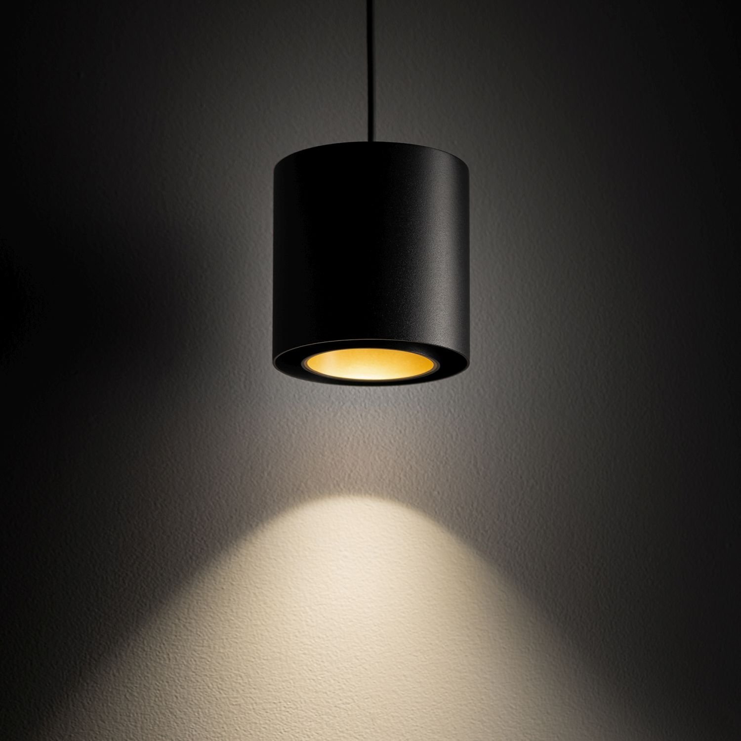 Czarna, cylindryczna lampa wisząca emituje ciepłe, żółte światło na ścianę. Lampa jest zawieszona na cienkim przewodzie.
