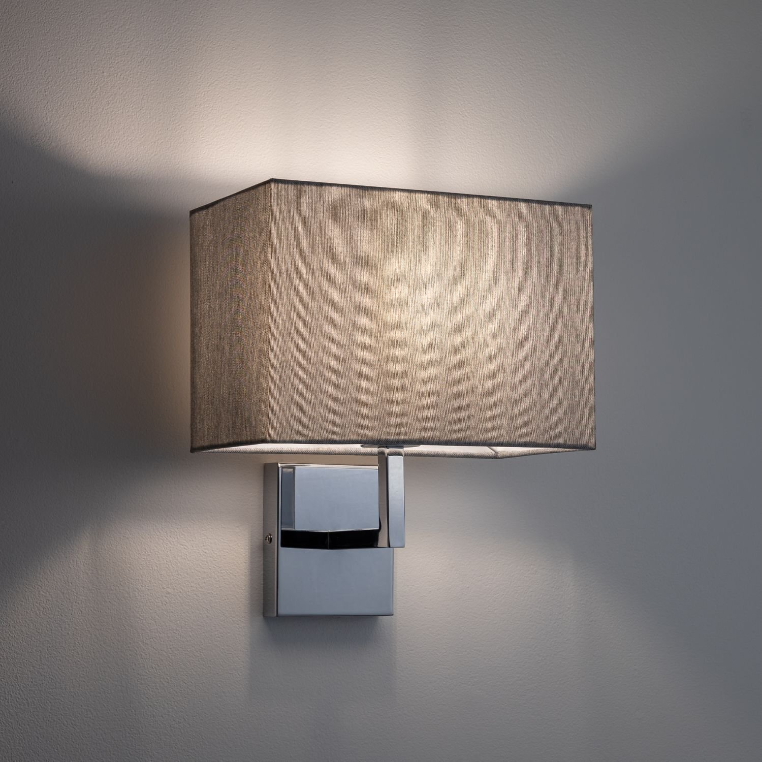 Na obrazie widoczny jest kinkiet z prostokątnym abażurem w kolorze szarym, zamontowany na ścianie. Podstawa lampy jest metaliczna i ma prosty, nowoczesny design.