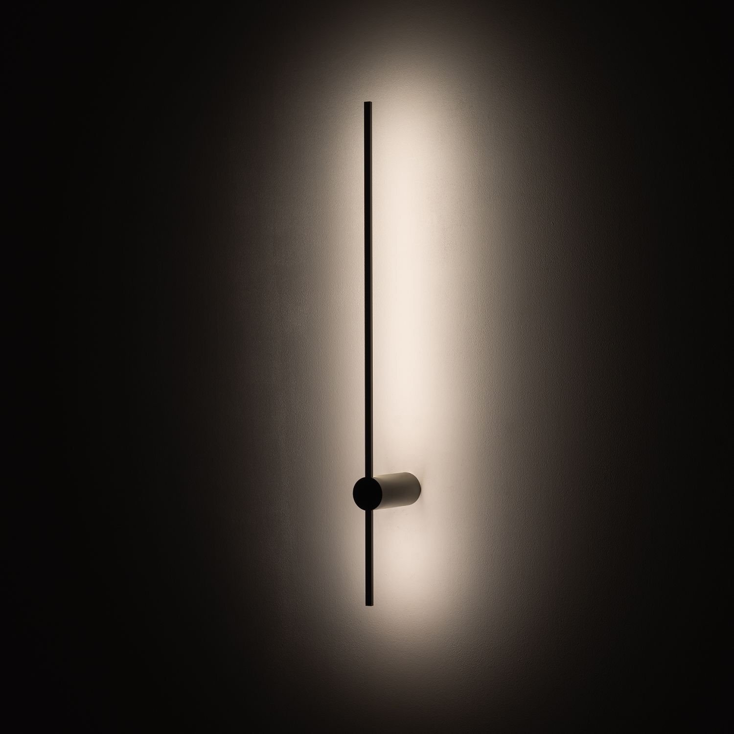 Na obrazie widoczna jest nowoczesna lampa ścienna o prostym, pionowym kształcie, emitująca delikatne światło na ścianę. Lampa ma minimalistyczny design z ciemnym korpusem i jasnym oświetleniem.
