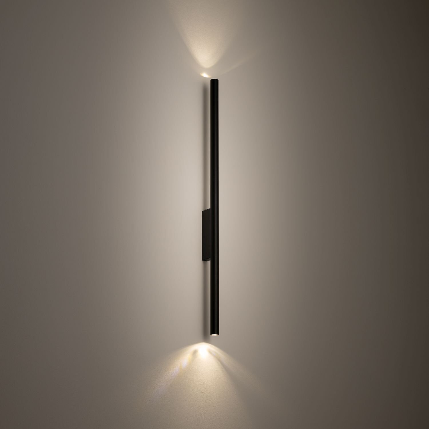 Na obrazie widoczna jest nowoczesna lampa ścienna w kształcie pionowego, czarnego pręta emitującego światło na górze i na dole. Lampa jest zamontowana na jasnej ścianie, tworząc delikatne efekty świetlne.