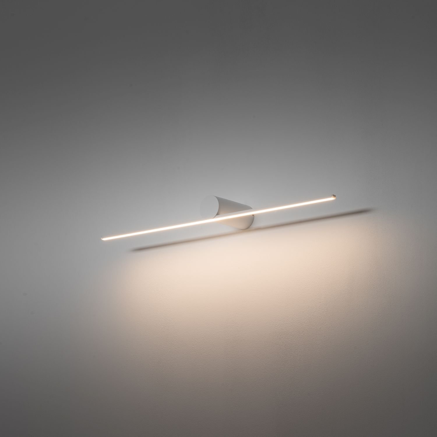 Na obrazie widoczna jest nowoczesna lampa ścienna LED o prostym, minimalistycznym designie, emitująca delikatne światło na ścianę. Lampa ma kształt długiego, cienkiego pręta zamocowanego na stożkowej podstawie.