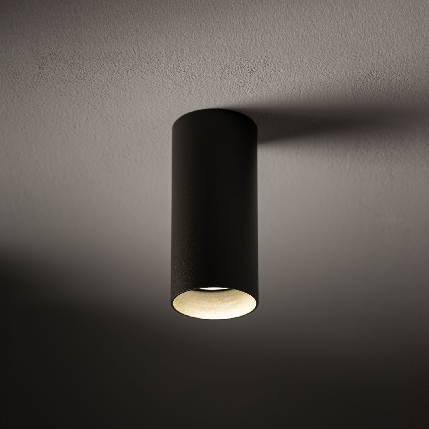 Cylindryczna, czarna lampa natynkowa zamontowana na suficie, emitująca światło w dół. Powierzchnia sufitu jest gładka i jasna.