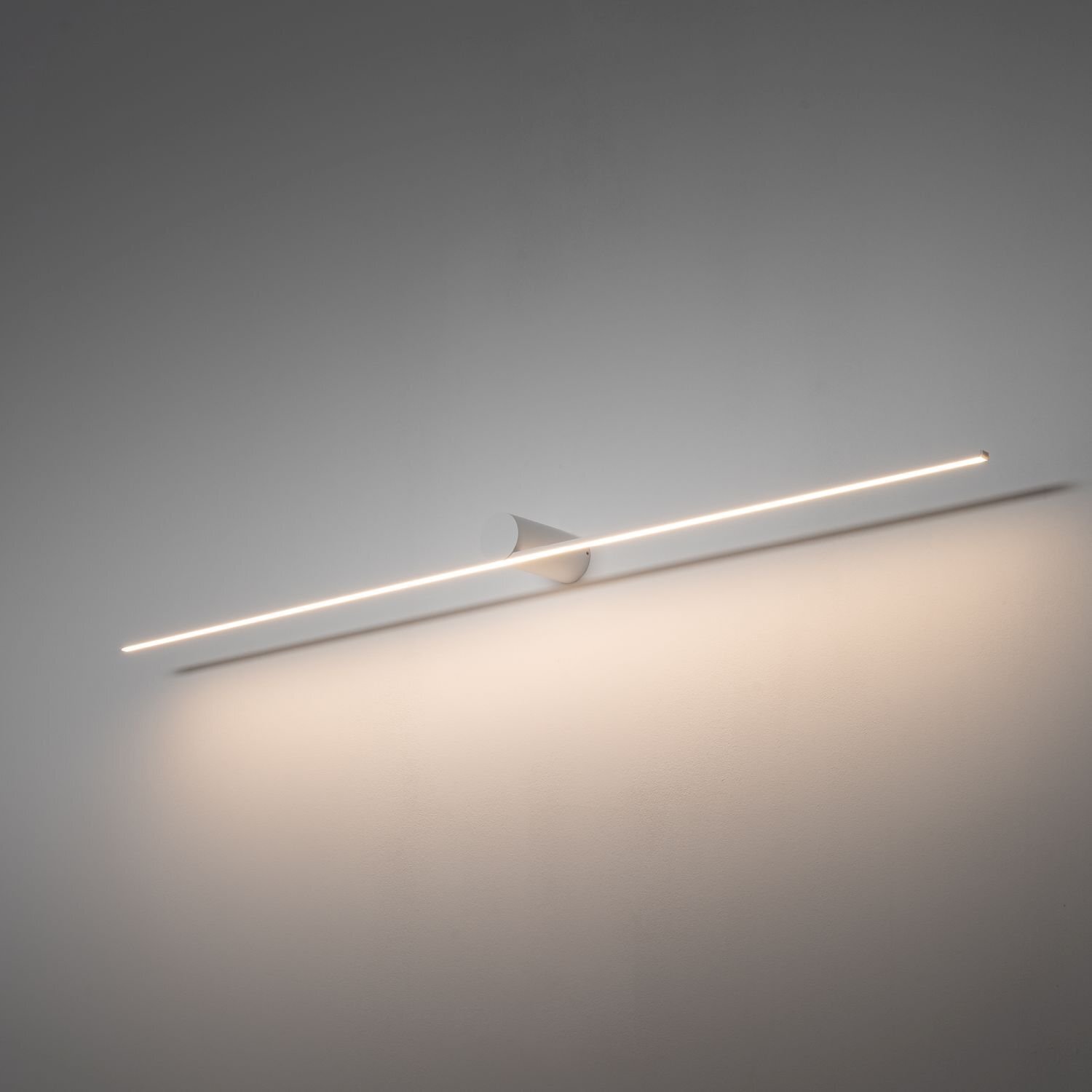 Na obrazie widoczna jest nowoczesna lampa ścienna LED o prostym, liniowym kształcie, emitująca delikatne światło na ścianę. Lampa jest zamontowana na gładkiej, jasnej powierzchni.