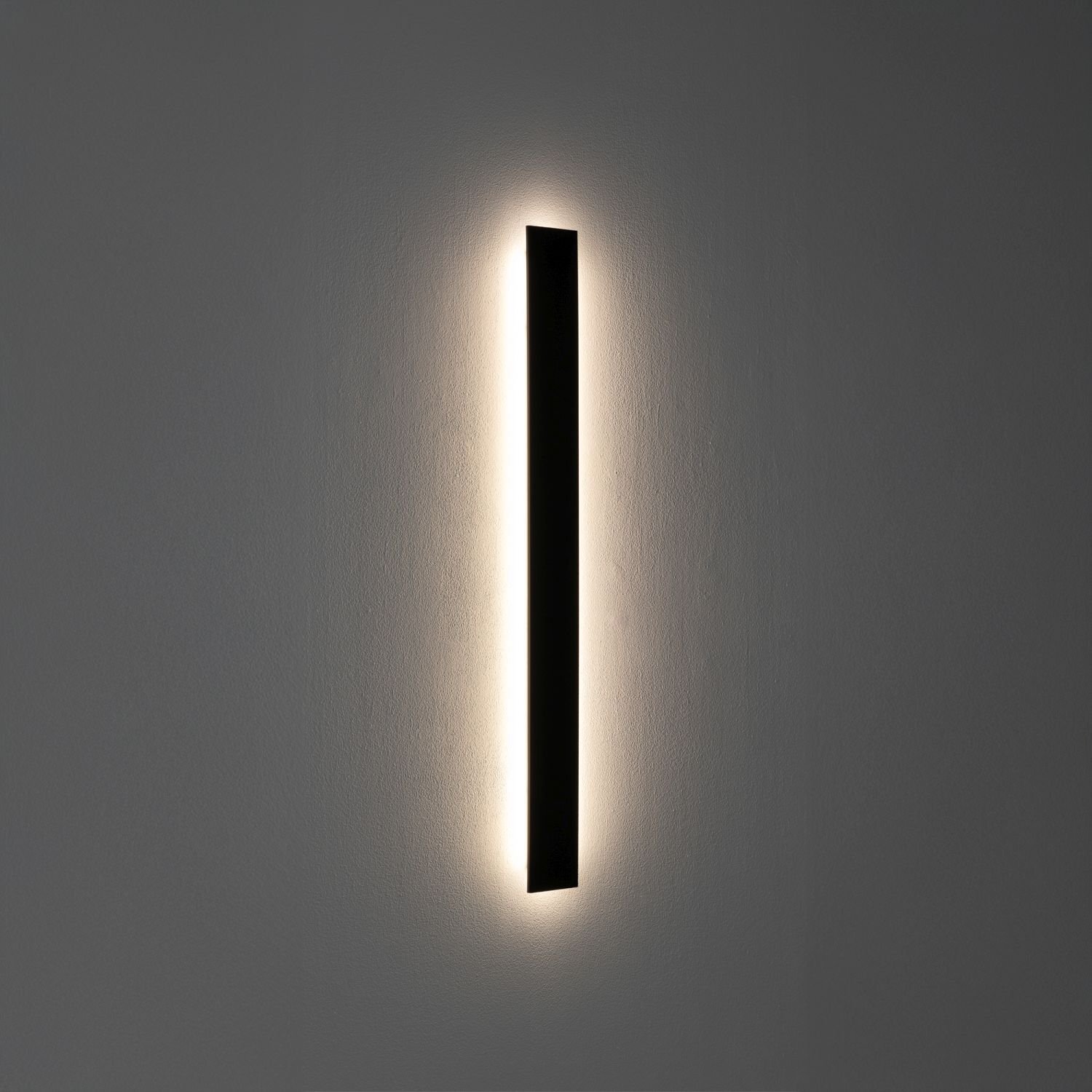 Na obrazie widoczna jest nowoczesna, liniowa lampa ścienna LED emitująca delikatne światło wokół ciemnego, pionowego panelu. Lampa jest zamontowana na gładkiej, jasnej ścianie.
