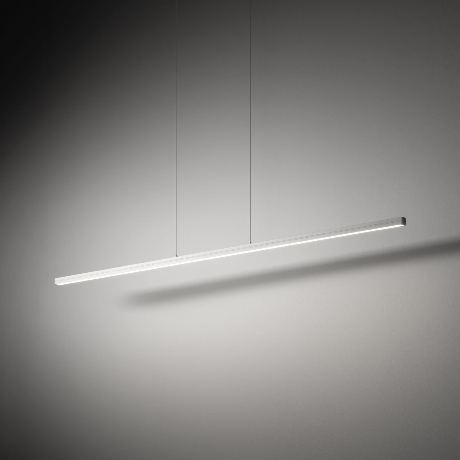 Na obrazie widoczna jest nowoczesna, wisząca lampa LED o prostym, liniowym kształcie. Lampa jest zawieszona na dwóch cienkich przewodach na tle jednolitej, jasnej ściany.