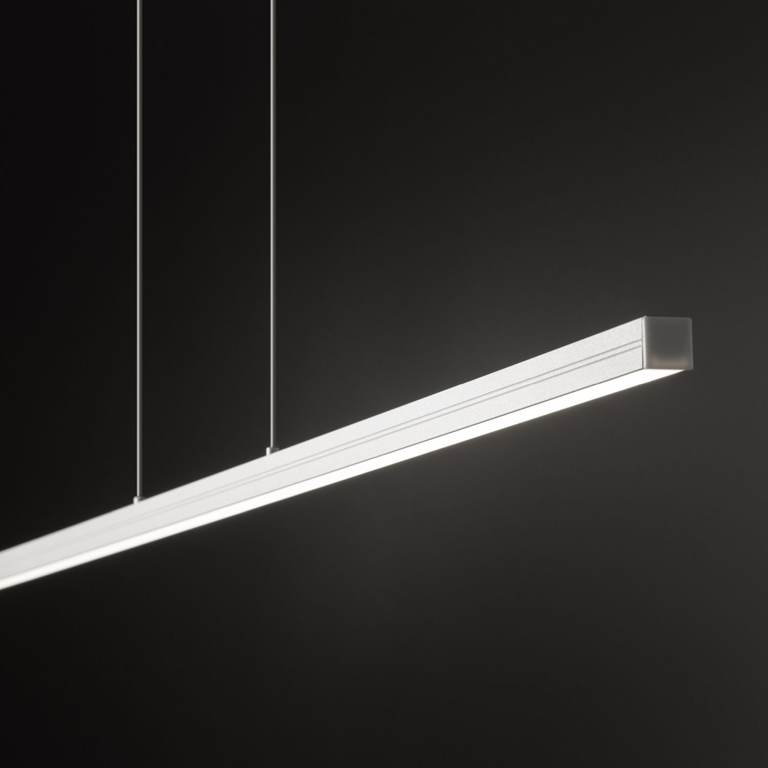 Na obrazie widoczna jest nowoczesna lampa wisząca o prostokątnym kształcie, zawieszona na dwóch cienkich linkach. Lampa emituje jasne, białe światło na ciemnym tle.