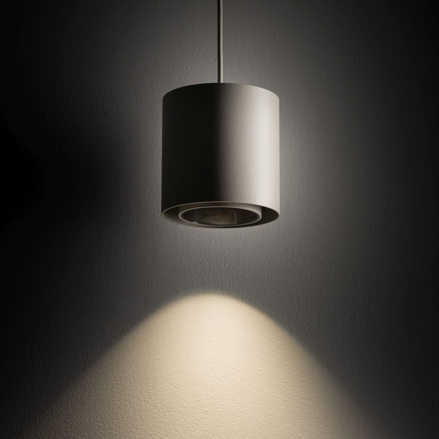 Na obrazie widoczna jest nowoczesna, cylindryczna lampa wisząca emitująca światło na ścianę, tworząc jasny okrąg na powierzchni. Lampa ma gładką, matową powierzchnię i jest zawieszona na cienkim przewodzie.