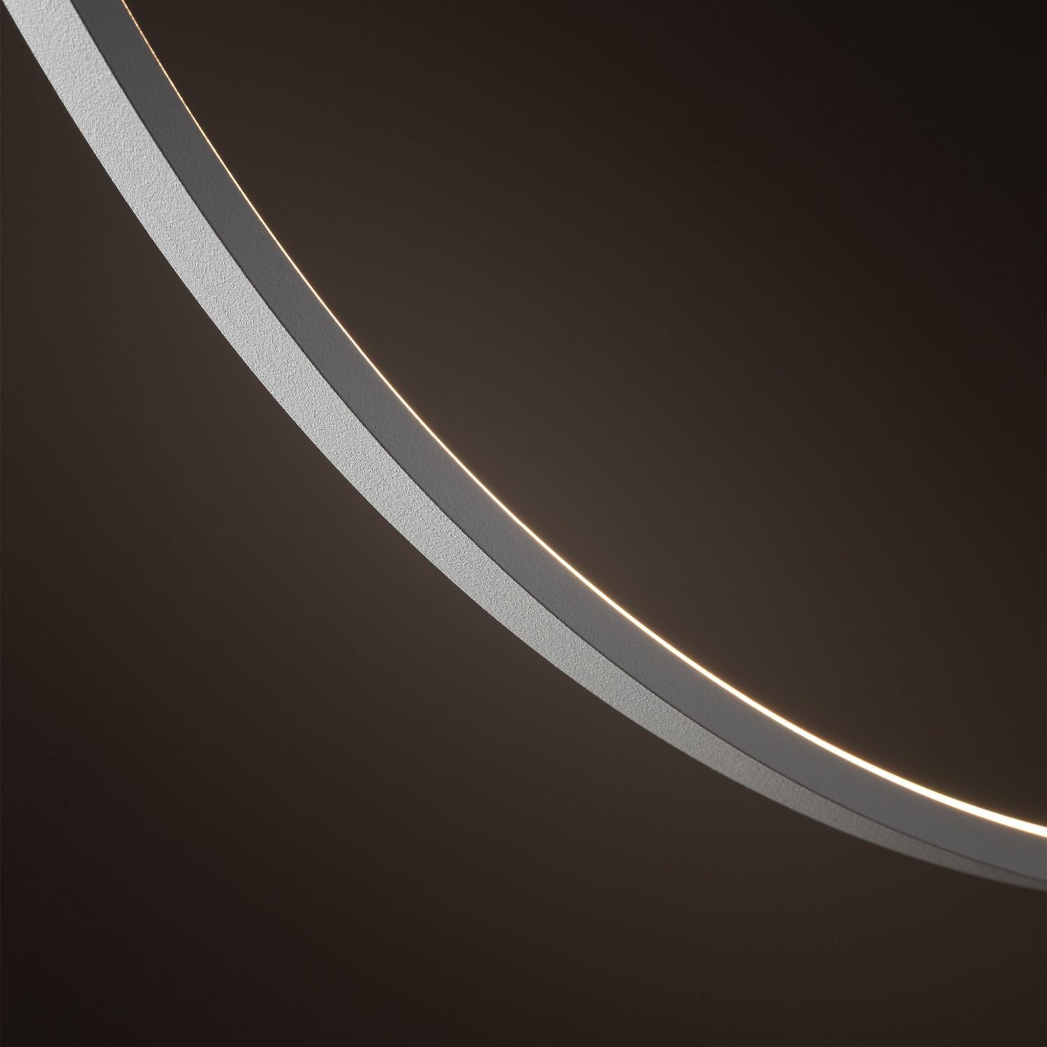 Obraz przedstawia fragment okrągłej lampy wiszącej z delikatnym, białym światłem LED na ciemnym tle. Lampa ma nowoczesny design z gładką, metaliczną powierzchnią.