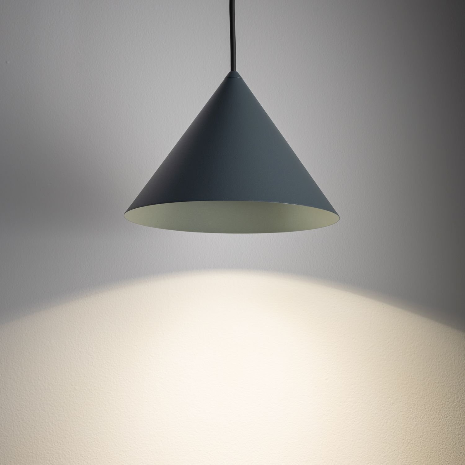 Na obrazie widoczna jest wisząca lampa o stożkowym kształcie, emitująca światło na ścianę. Lampa ma ciemny kolor z zewnątrz i jasne wnętrze.