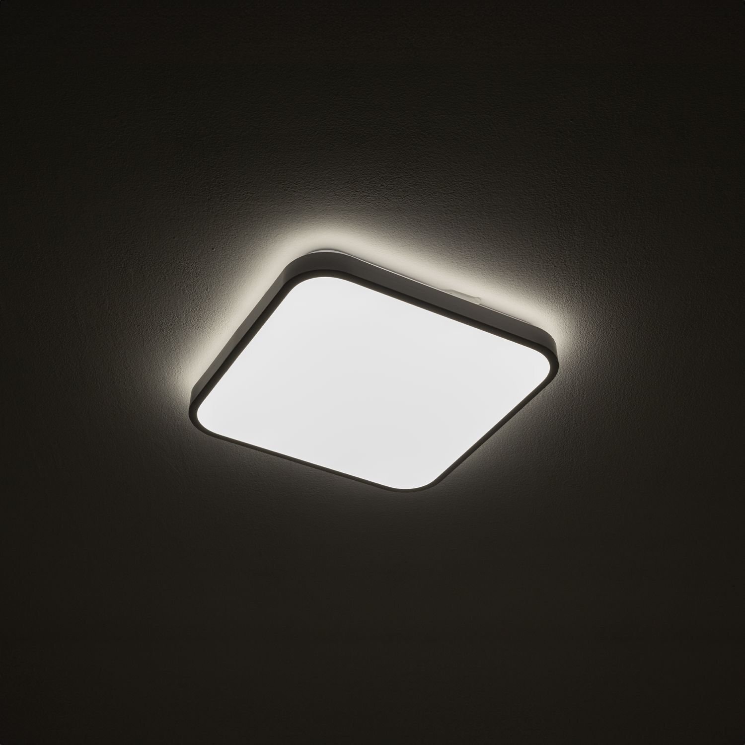 Kwadratowy plafon LED emituje jasne światło, tworząc delikatną poświatę na suficie. Lampa ma nowoczesny design i jest zamontowana na ciemnym tle.