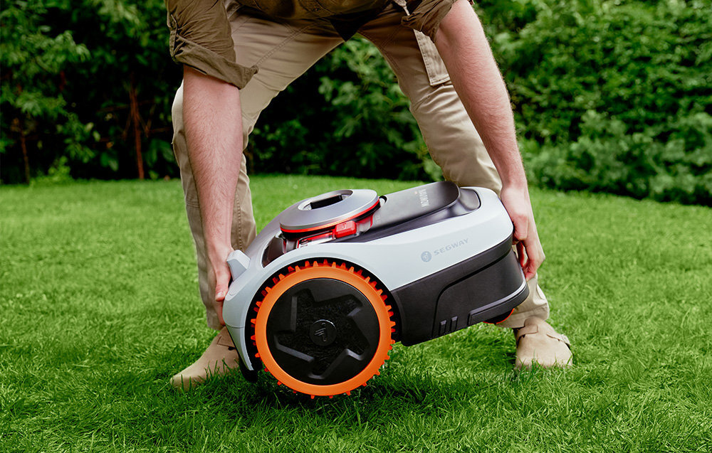 Osoba trzyma robot koszący z widocznym logo SEGWAY, stojący na trawie. Robot ma pomarańczowe koła i szaro-czarną obudowę.