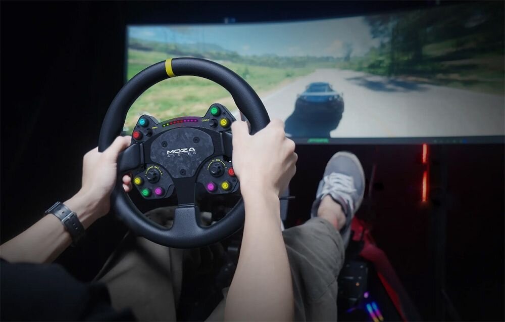 Osoba trzyma kierownicę z napisem \'MOZA Racing\', przed dużym ekranem z widokiem na grę wyścigową.