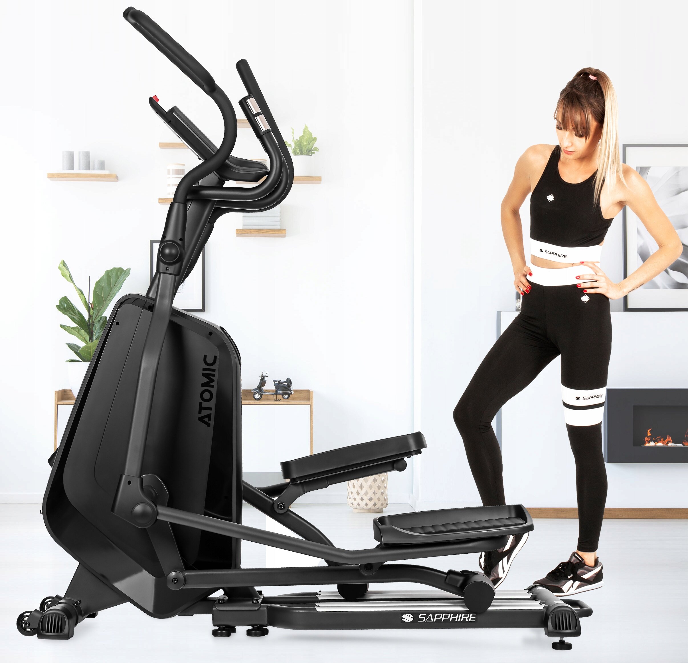 Na obrazie znajduje się czarny orbitrek z napisem \'ATOMIC\' na obudowie oraz \'SAPPHIRE\' na podstawie. Obok stoi osoba ubrana w czarny strój sportowy z białymi elementami.