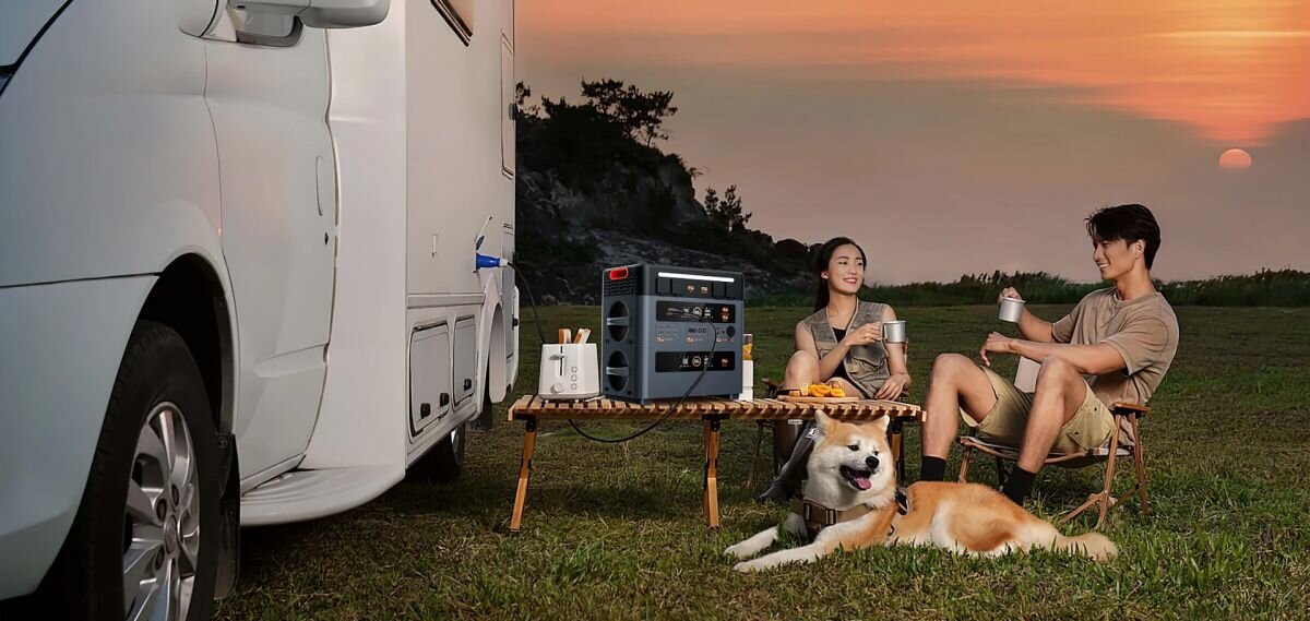 Stacja zasilania OSCAL PowerMax 2400 Pro 2016Wh, 12V, 24V, kamper, zasilanie, PowerMax 2400 Pro na stoliku przy kamperze, z dwiema osobami i psem na trawie.
