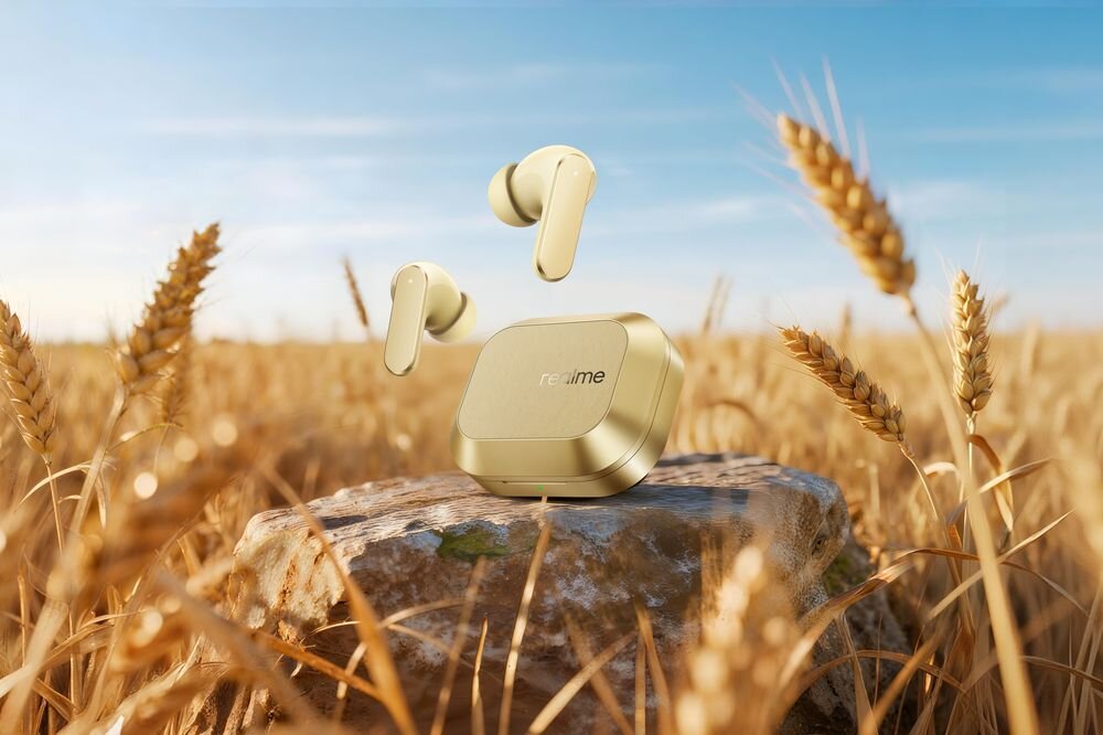 Złote słuchawki dokanałowe i etui ładujące leżą na kamieniu w polu pszenicy. Na etui widoczne jest logo \'realme\'.