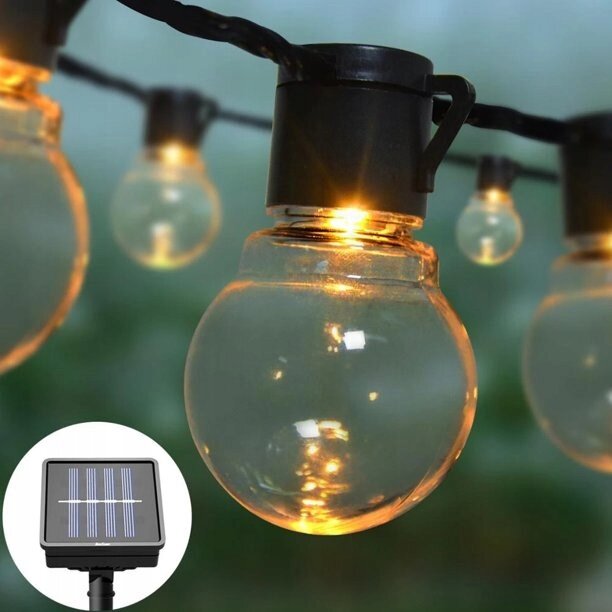 Na obrazie widoczne są przezroczyste żarówki wiszące na czarnym kablu, emitujące ciepłe światło. W rogu znajduje się mały panel solarny z widocznymi paskami ogniw.