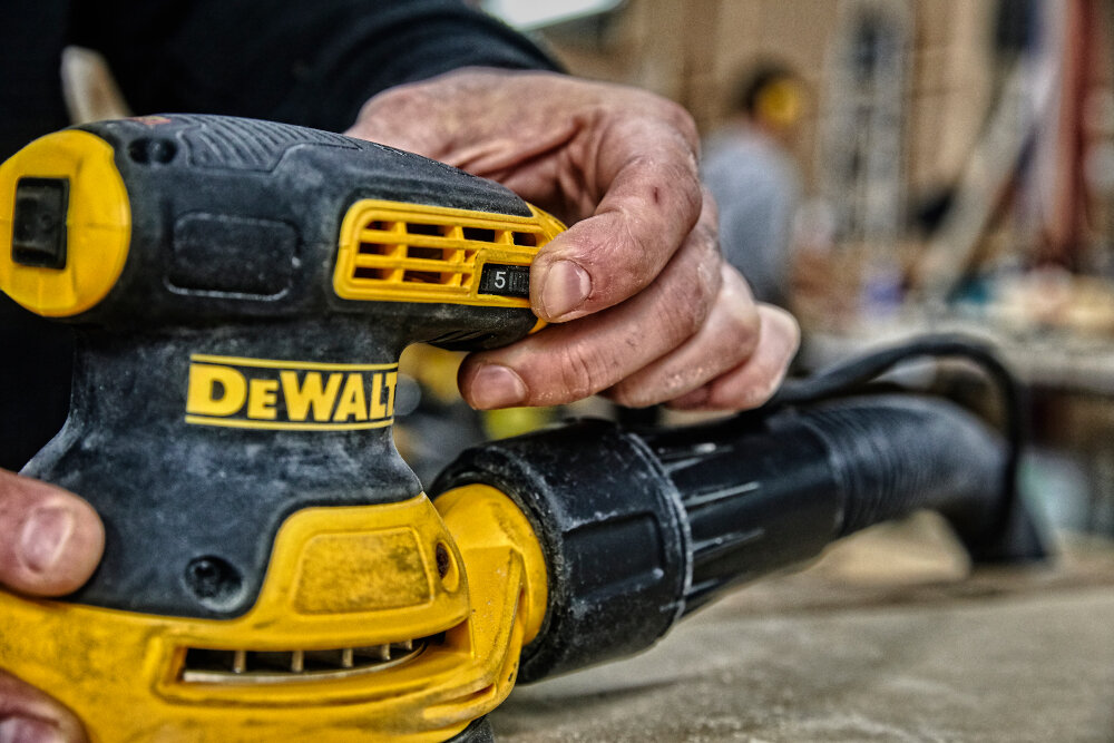 Osoba trzyma szlifierkę mimośrodową z widocznym logo DEWALT. Urządzenie jest czarno-żółte, a w tle widać rozmyte elementy warsztatu.