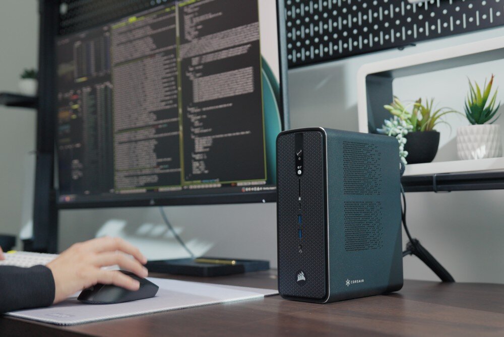Komputer CORSAIR AI Workstation 300 Komputer stacjonarny w ciemnej obudowie stojący pod kątem na biurku obok monitora wyświetlającego linie kodu, na pierwszym planie widoczna dłoń na czarnej myszce komputerowej, przeznaczenie, podzespoły, cechy