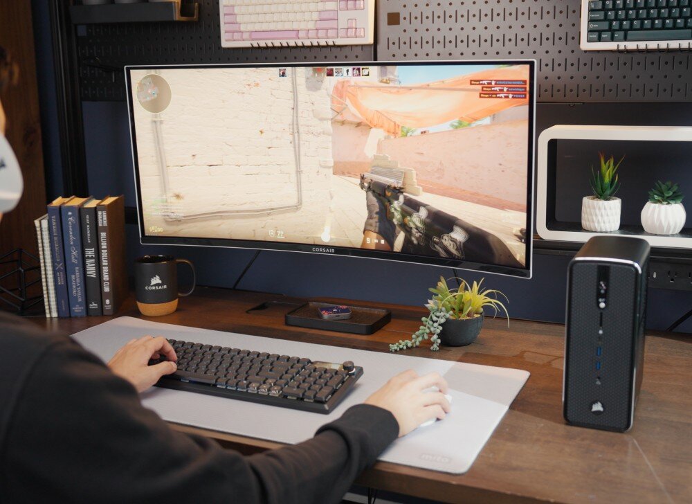 Komputer CORSAIR AI Workstation 300 Szerokie ujęcie stanowiska pracy z zakrzywionym monitorem panoramicznym wyświetlającym grę pierwszoosobową, po prawej stronie stoi mały komputer, a po lewej ułożone są książki i kubek, układ graficzny, pamięć