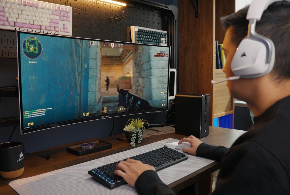 Komputer CORSAIR AI Workstation 300 Mężczyzna w białych słuchawkach nausznych siedzi tyłem do obiektywu przed dużym zakrzywionym monitorem z uruchomioną grą wideo, po prawej stronie ekranu na biurku stoi niewielki czarny komputer, dysk, pojemność, zasilacz