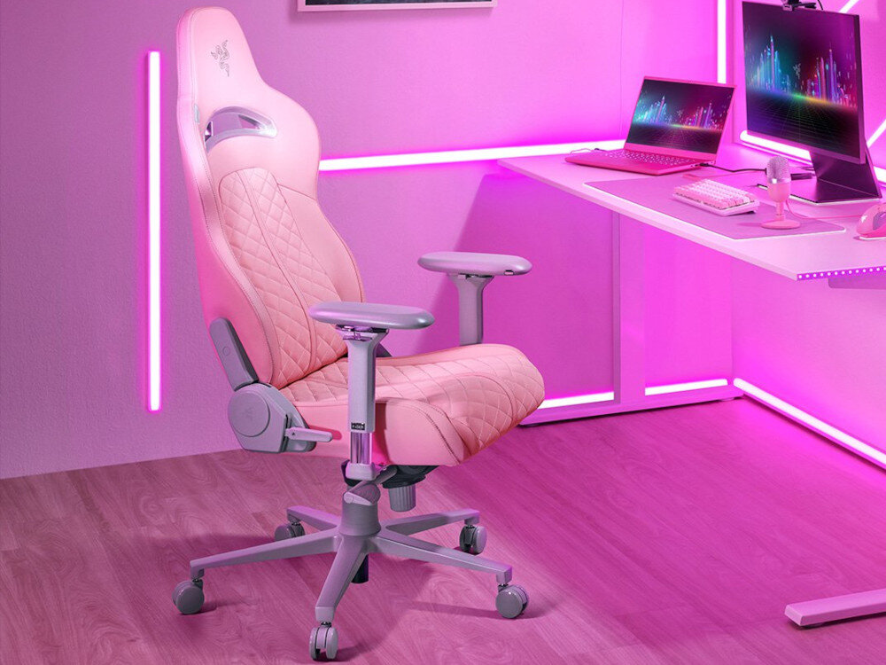 Różowy fotel gamingowy z logo Razer, ustawiony w nowoczesnym pokoju z różowym oświetleniem LED. Obok znajduje się biurko z laptopem, monitorem i klawiaturą w podobnej kolorystyce.
