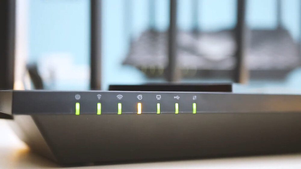 Na obrazie widoczny jest router z sześcioma świecącymi diodami LED na przednim panelu, z których jedna świeci na pomarańczowo, a pozostałe na zielono. W tle widać rozmyte elementy konstrukcji routera.