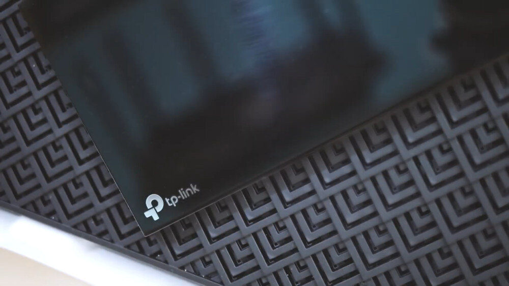 Obraz przedstawia fragment urządzenia z logo \'tp-link\' na błyszczącej powierzchni. W tle widoczna jest struktura złożona z powtarzających się wzorów geometrycznych.