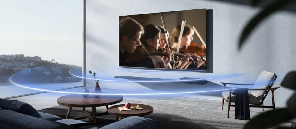 W nowoczesnym salonie na ścianie wisi duży telewizor, na którym wyświetlany jest obraz orkiestry smyczkowej. Wokół telewizora widoczne są efekty dźwiękowe w postaci niebieskich fal.