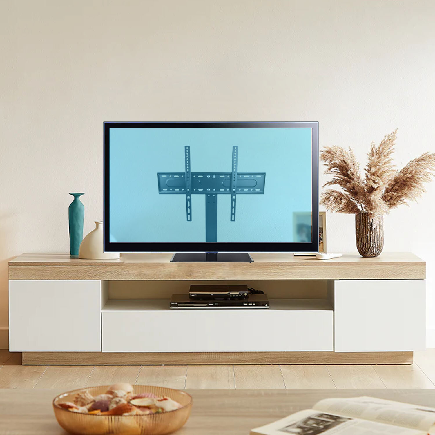 Na ekranie telewizora widoczny jest uchwyt do telewizora z pionowymi i poziomymi elementami montażowymi. Telewizor stoi na drewnianej szafce, obok której znajdują się dekoracyjne wazony i suszone trawy.