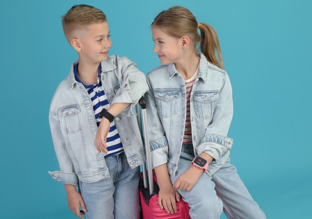 Dwoje dzieci siedzi na różowej walizce, każde z nich ma na sobie jasną kurtkę jeansową i smartwatch na nadgarstku. Tło jest jednolite, niebieskie.