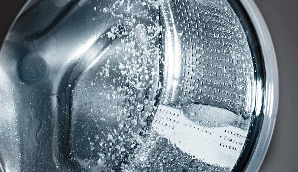 Zbliżenie na bęben pralki z widocznymi kroplami wody na metalowej powierzchni. Wnętrze bębna jest oświetlone, co podkreśla jego teksturę i połysk.