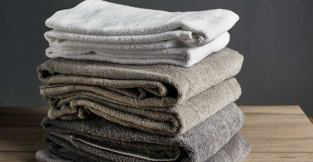 Stos złożonych ręczników w różnych odcieniach szarości i bieli leży na drewnianej powierzchni. Tło jest jednolite i ciemne.