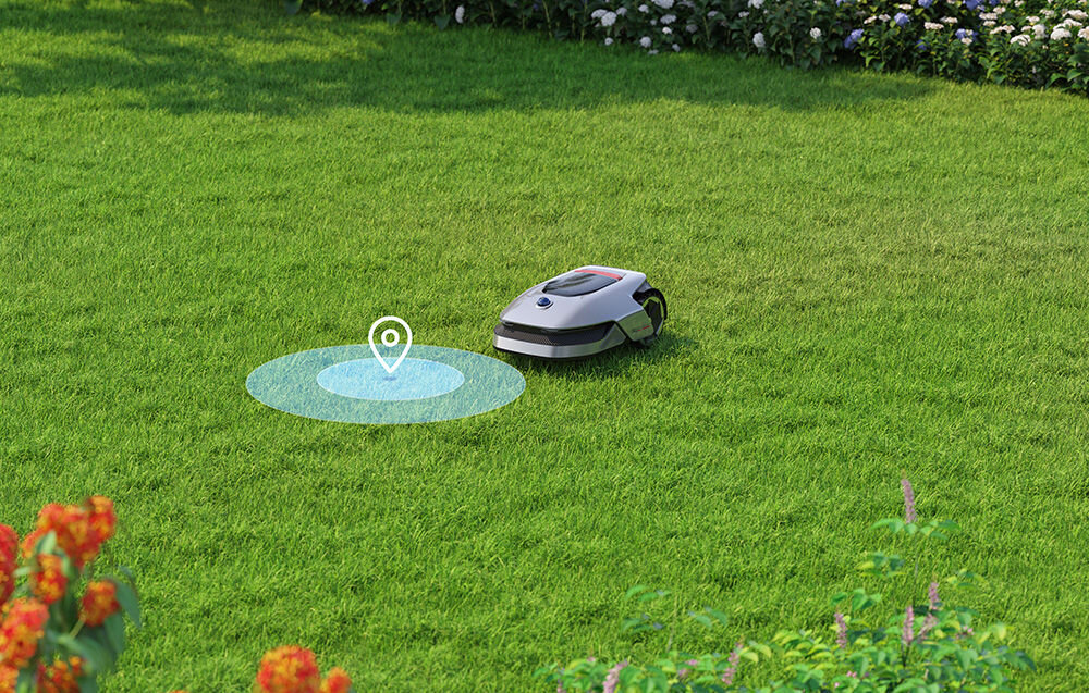 Na trawniku znajduje się robot koszący z widocznym symbolem lokalizacji nad trawą. W tle widać kwiaty i krzewy.