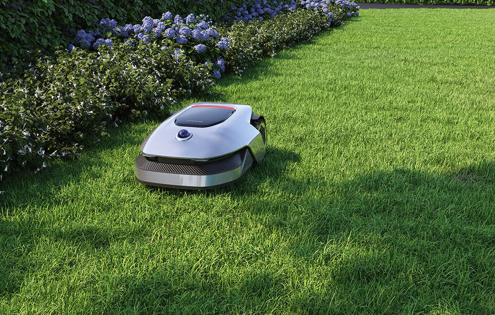 Na obrazie widoczny jest robot koszący trawę na zielonym trawniku, otoczony żywopłotem z kwiatami. Robot ma nowoczesny, srebrny design z ciemnymi elementami.