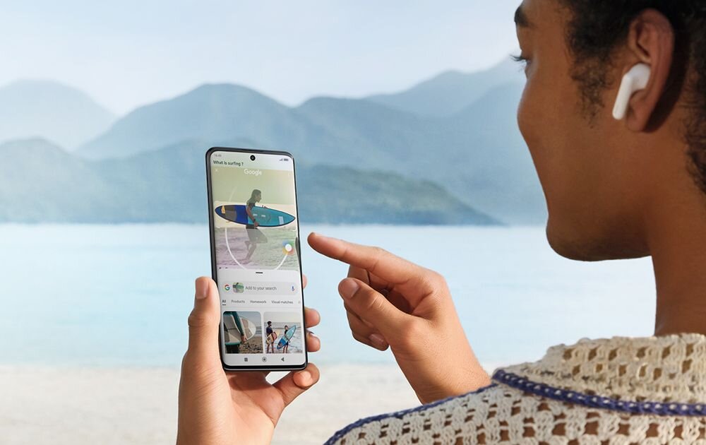 Osoba trzyma smartfon, na ekranie widoczna jest aplikacja z obrazem osoby z deską surfingową. W tle znajduje się jezioro i góry.
