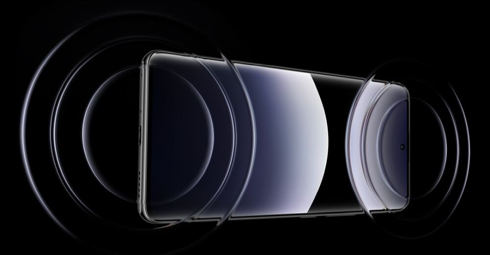 Obraz przedstawia smartfon z widocznym ekranem i obudową w ciemnym otoczeniu. Wokół urządzenia widoczne są koncentryczne okręgi, sugerujące efekt dźwiękowy lub wizualny.