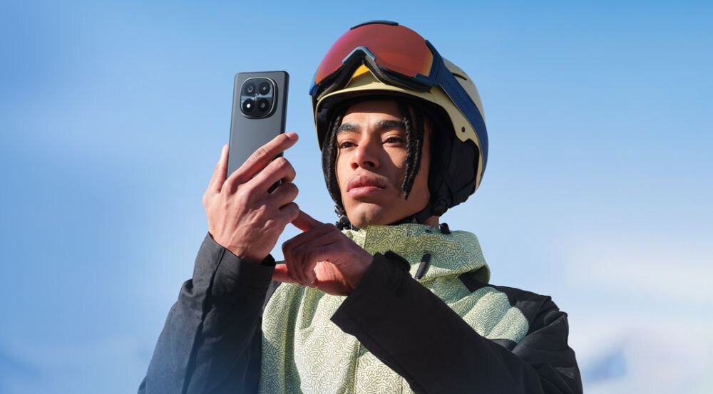 Osoba w kasku narciarskim trzyma smartfon, patrząc na jego ekran. W tle widać jasne, niebieskie niebo.