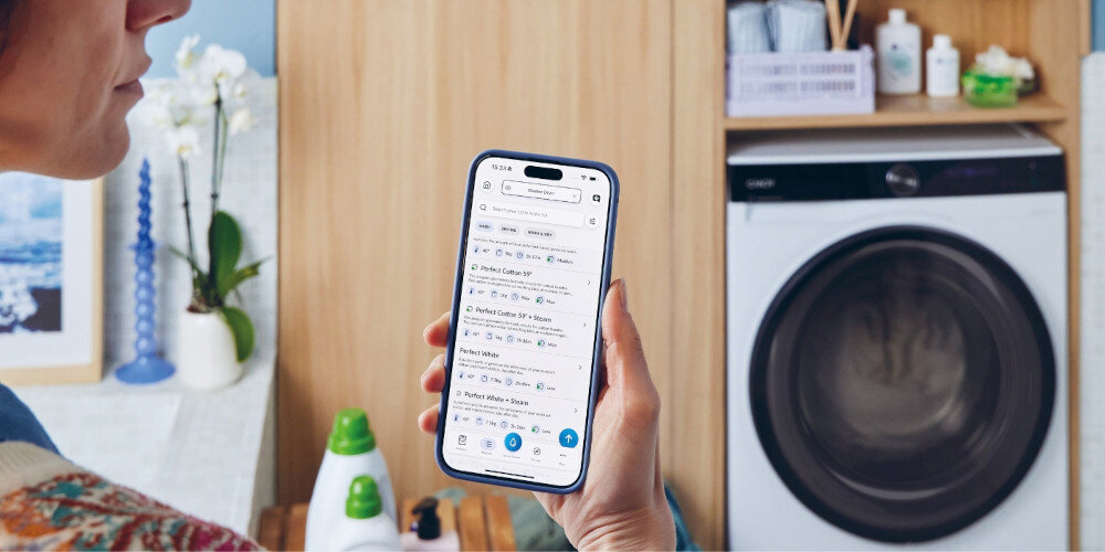 Osoba trzyma smartfon z aplikacją do zarządzania pralko-suszarką, w tle widoczna jest pralko-suszarka CANDY z dużym okrągłym bębnem. Na półce obok znajdują się różne akcesoria do prania.