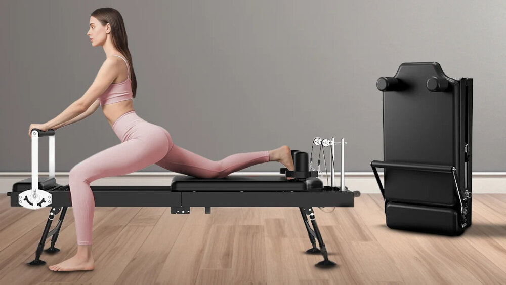 Osoba ćwiczy na czarnym reformerze do pilatesu, ubrana w różowy strój sportowy. Obok znajduje się złożony reformer w pozycji pionowej.