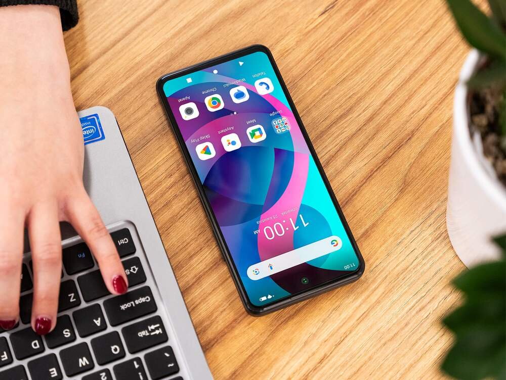 Na drewnianym stole leży smartfon z kolorowym wyświetlaczem, na którym widoczne są ikony aplikacji i godzina 11:00. Obok znajduje się ręka na klawiaturze laptopa z naklejką Intel Inside.