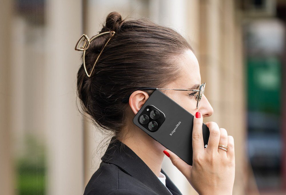 Osoba trzyma przy uchu czarny smartfon z widocznym logo \'Kruger&Matz\'.