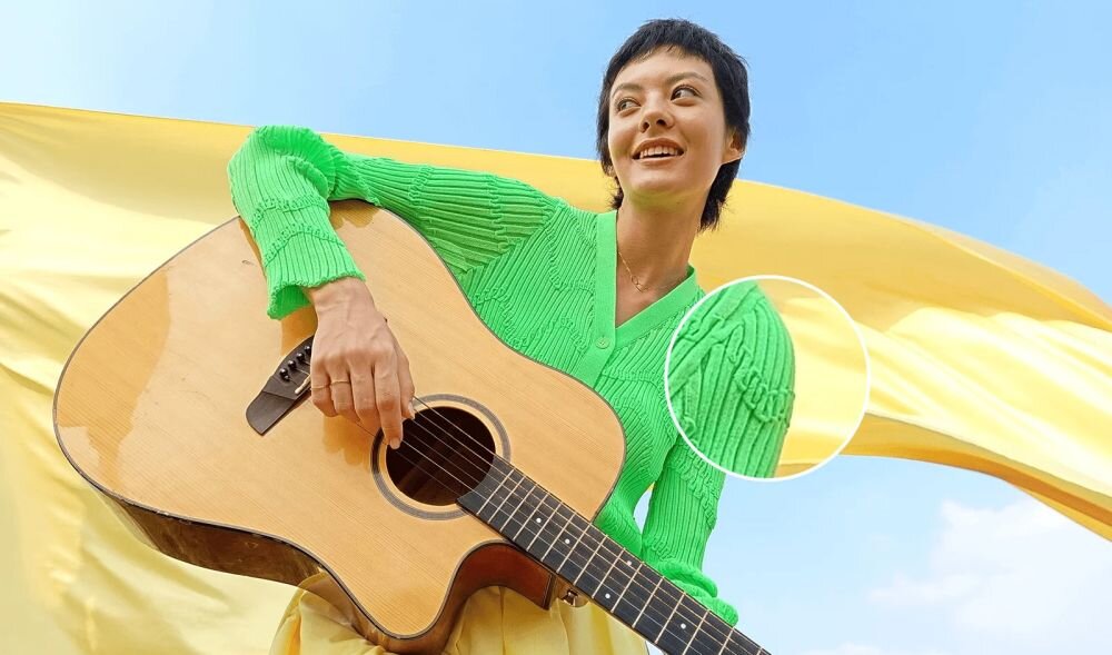 Osoba w zielonym swetrze trzyma gitarę akustyczną na tle żółtej tkaniny i błękitnego nieba. Na swetrze widoczny jest wzór w postaci linii i warkoczy.