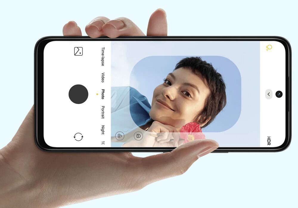 Osoba trzyma smartfon, na ekranie którego widać aplikację aparatu z włączonym trybem \'Photo\'. Na ekranie wyświetla się zdjęcie osoby z krótkimi włosami, uśmiechającej się do kamery.