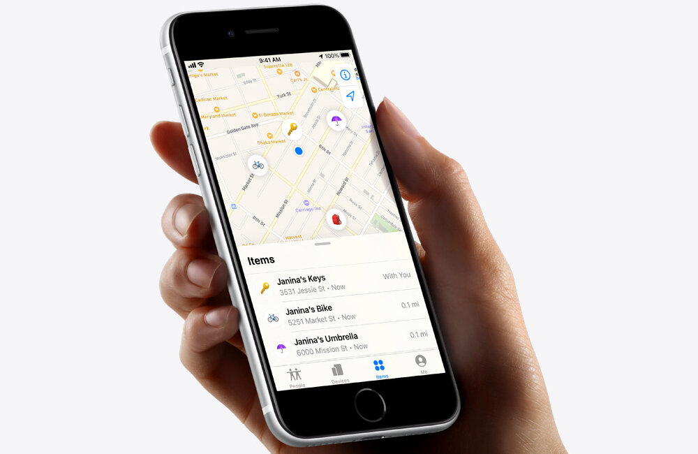 Osoba trzyma smartfon z aplikacją mapową, na której widoczne są lokalizacje przedmiotów: Janina\'s Keys, Janina\'s Bike i Janina\'s Umbrella. Na ekranie widać mapę z ikonami oraz listę przedmiotów z ich lokalizacjami i odległościami.