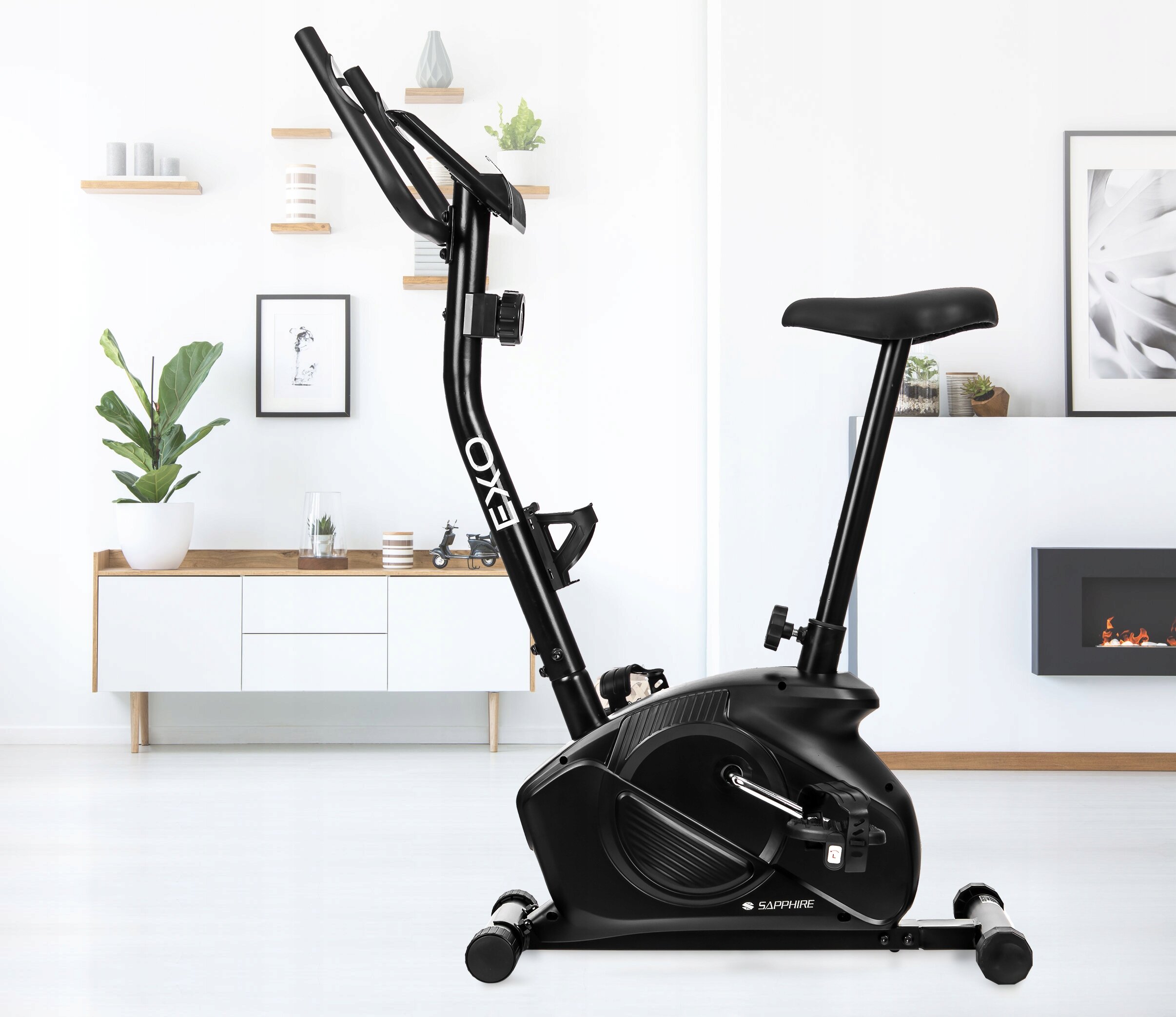 Czarny rower treningowy stacjonarny magnetyczny pionowy z napisem \'EXO\' na ramie, ustawiony w nowoczesnym wnętrzu z białymi ścianami i drewnianymi półkami. W tle widoczna jest komoda z rośliną i dekoracjami oraz kominek.