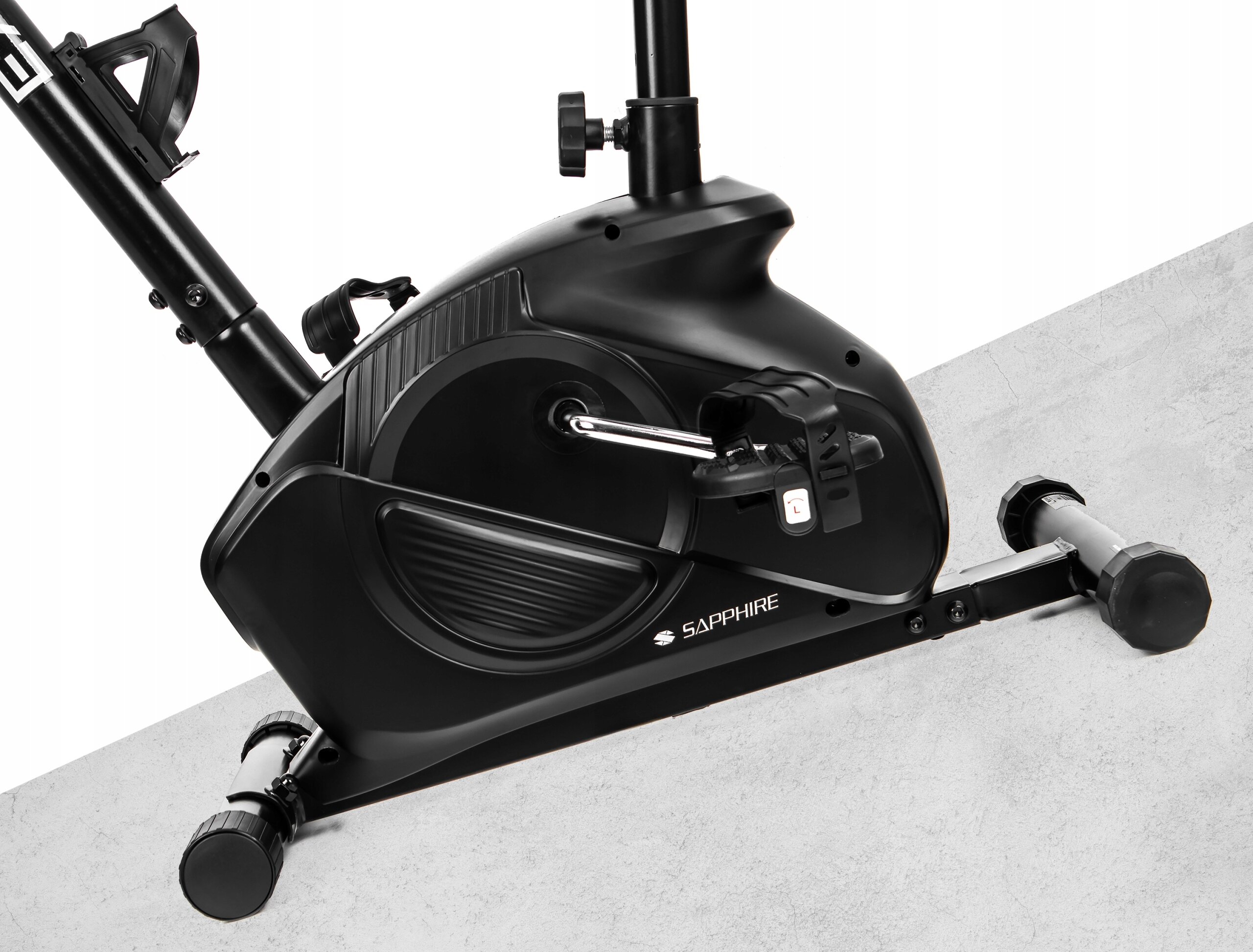 Czarny rower treningowy stacjonarny z widocznym pedałem i napisem \'SAPPHIRE\' na obudowie. Urządzenie stoi na jasnej powierzchni.