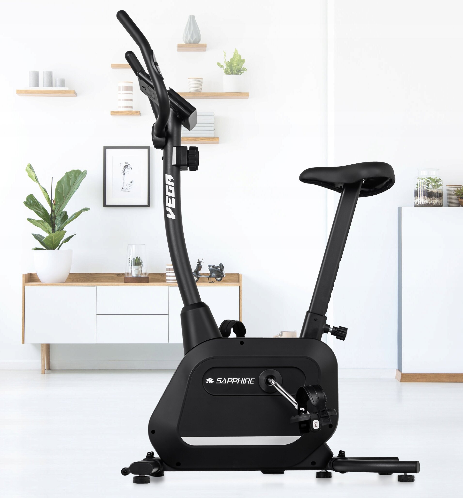 Czarny rower treningowy stacjonarny z napisem \'VEGA\' na kierownicy i \'SAPPHIRE\' na obudowie. W tle jasne wnętrze z białymi meblami i roślinami.