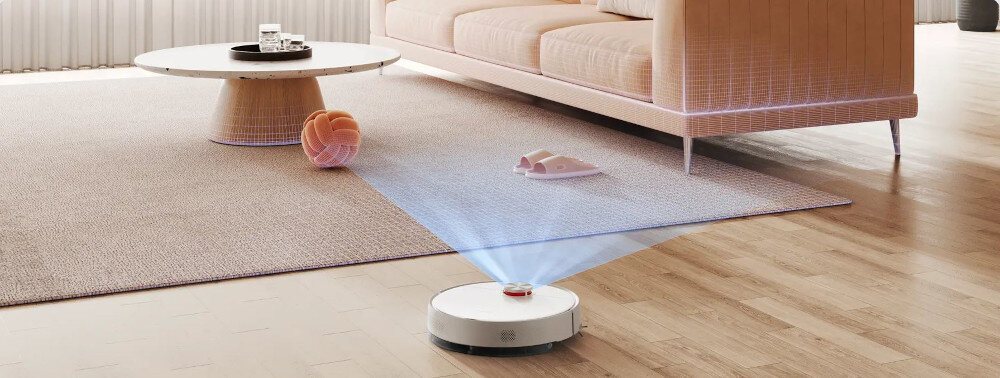 Na drewnianej podłodze w salonie znajduje się biały robot sprzątający z niebieskim światłem skierowanym w stronę dywanu. W tle widać stolik kawowy i sofę.